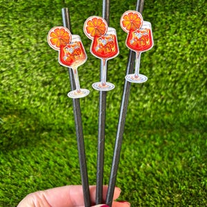 May include: Three black paper straws decorated with cocktail stickers. Each sticker shows a red cocktail in a stemmed glass, topped with an orange slice. The straws are set against a green background.