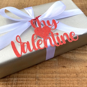 Puede incluir: Una caja de regalo plateada con una cinta de sat&eacute;n blanca y una etiqueta de papel con purpurina roja que dice "My Valentine".