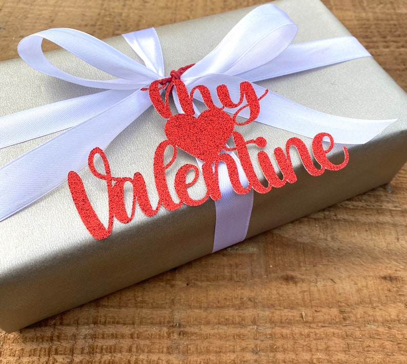 Puede incluir: Una caja de regalo plateada con una cinta de sat&eacute;n blanca y una etiqueta de papel con purpurina roja que dice "My Valentine".