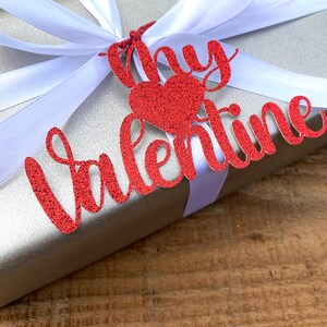 Puede incluir: Una caja de regalo plateada con una cinta blanca y una etiqueta roja con purpurina que dice "My Valentine" con un coraz&oacute;n rojo con purpurina.