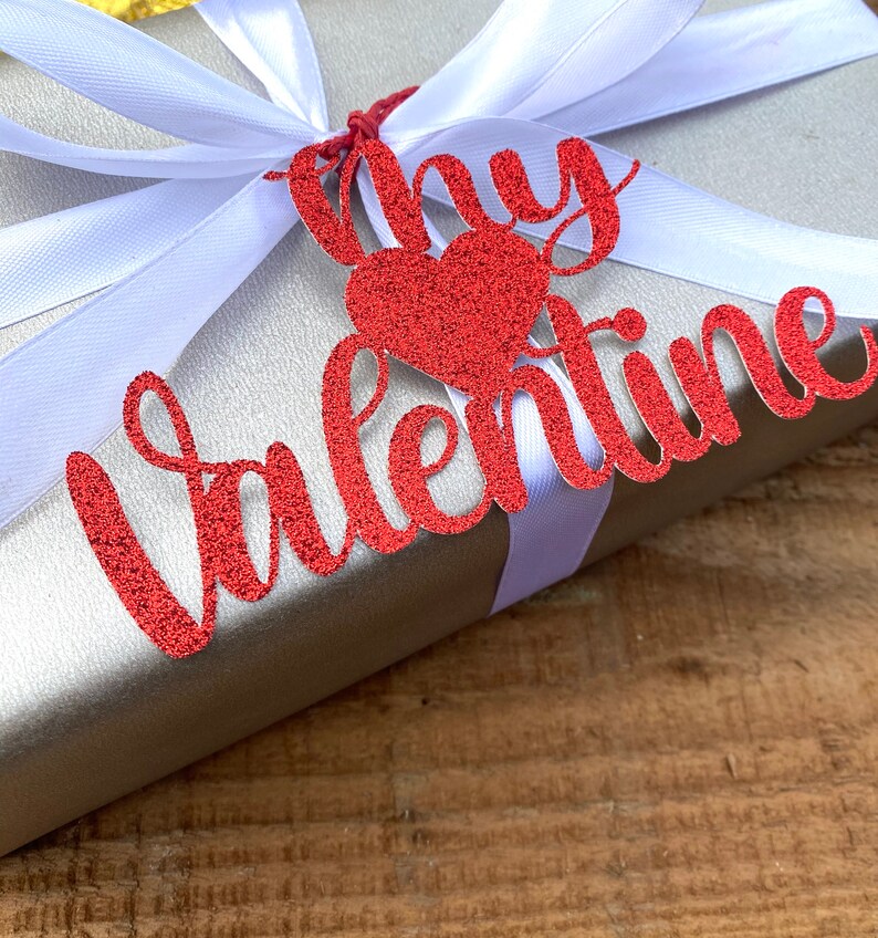 Puede incluir: Una caja de regalo plateada con una cinta blanca y una etiqueta roja con purpurina que dice "My Valentine" con un coraz&oacute;n rojo con purpurina.