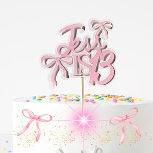 May include: A white iced cake decorated with colourful sprinkles and pink bow details. A pink glitter cake topper reads "Jess is 13". A pink bow design is also on the cake. A cupcake is in the background.