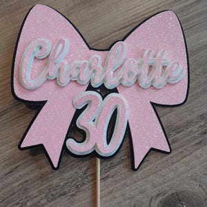 May include: A pink glitter bow-shaped cake topper with the name "Charlotte" and the number "30" in white and pink. The bow has a black outline and is mounted on a wooden stick.