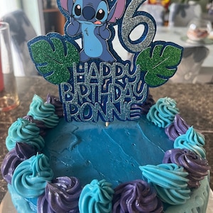K&ouml;nnte beinhalten: Ein runder Geburtstagskuchen mit blauer Glasur und dekorativen Wirbeln in Blau- und Lilat&ouml;nen. Ein Tortenaufsatz zeigt die Figur Stitch, die Zahl 6 und den Text "HAPPY BIRTHDAY RONNIE".