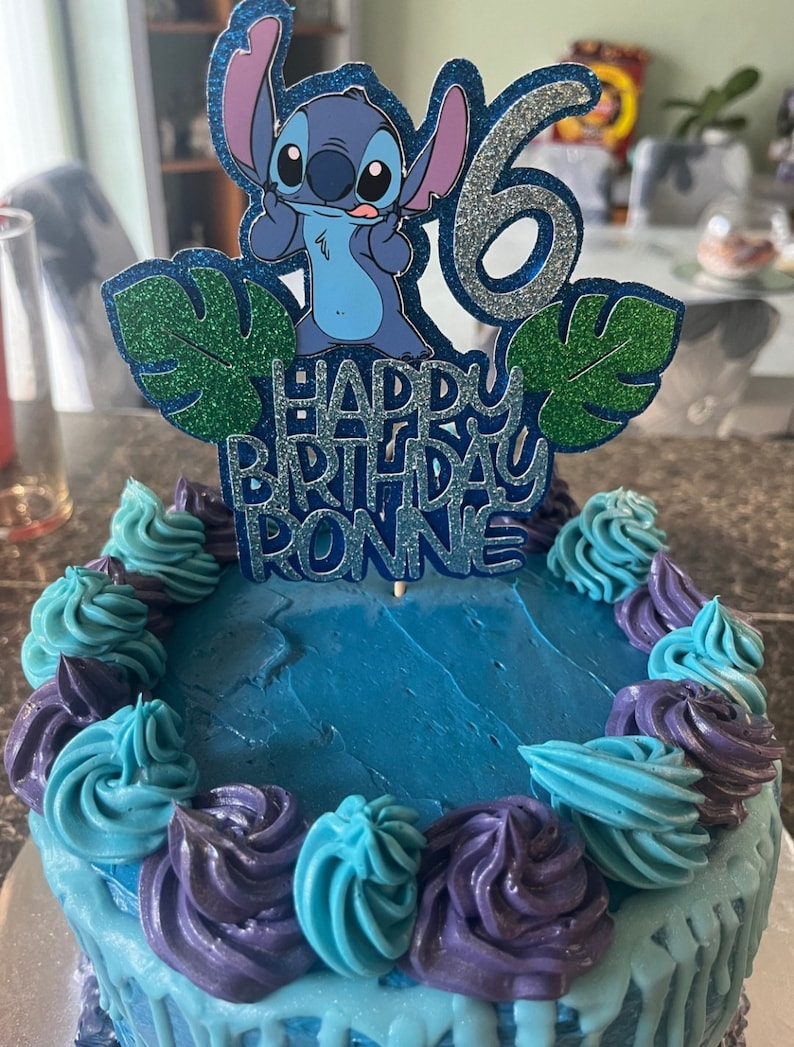 K&ouml;nnte beinhalten: Ein runder Geburtstagskuchen mit blauer Glasur und dekorativen Wirbeln in Blau- und Lilat&ouml;nen. Ein Tortenaufsatz zeigt die Figur Stitch, die Zahl 6 und den Text "HAPPY BIRTHDAY RONNIE".