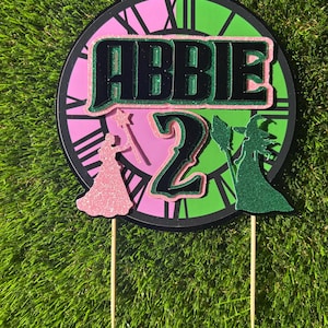 May include: A cake topper featuring the name "Abbie" and the number "2" in black, green, and pink glitter. The design includes silhouettes of a princess and a witch, set against a clock face. The topper is mounted on wooden sticks.