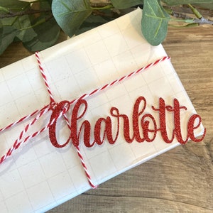 Puede incluir: Un regalo blanco envuelto con cuerda roja y blanca y una etiqueta de nombre de brillo rojo que dice "Charlotte".