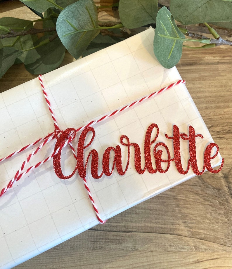 Puede incluir: Un regalo blanco envuelto con cuerda roja y blanca y una etiqueta de nombre de brillo rojo que dice "Charlotte".