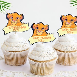 May include: Three cupcakes decorated with white icing and gold sprinkles. Each cupcake has a  "Happy Birthday Toby"  topper featuring a cartoon lion from The Lion King.
