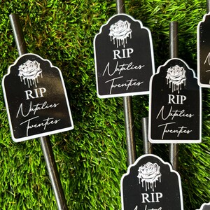 May include: Four black and white tombstone-shaped paper straws with a white rose and dripping paint design. The straws have the text "RIP" and "Naptlies Twenties" printed on them. The straws are displayed on a green artificial grass surface.