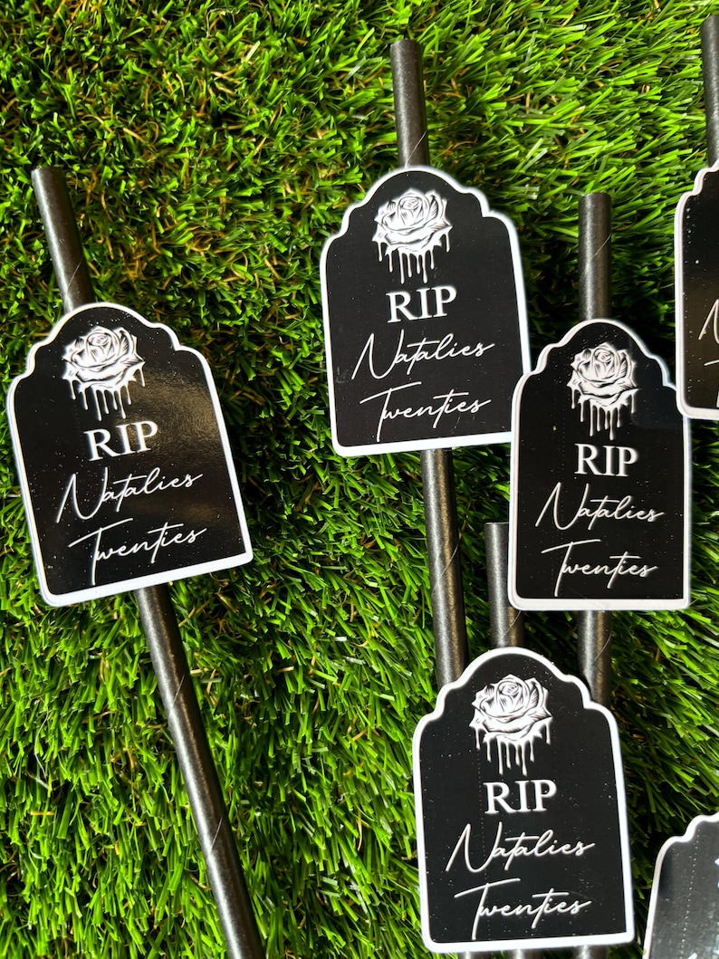 May include: Four black and white tombstone-shaped paper straws with a white rose and dripping paint design. The straws have the text "RIP" and "Naptlies Twenties" printed on them. The straws are displayed on a green artificial grass surface.