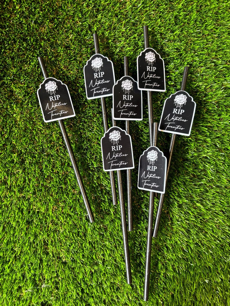 May include: Eight black straws with black and white paper flags attached. The flags have a white rose and the text "RIP Natalies Twenties".