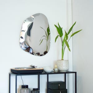 May include: A modern, irregularly shaped silver mirror hangs on a white wall. Below, a black metal table holds a record player, a white bowl, and a small decorative bottle. A potted plant with green leaves adds a touch of nature.