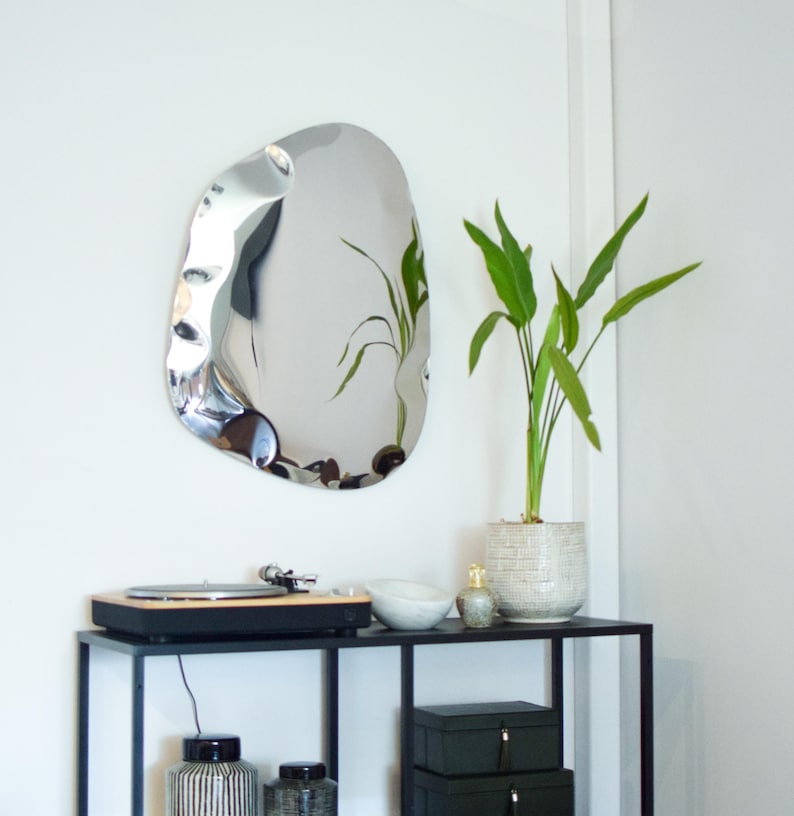 May include: A modern, irregularly shaped silver mirror hangs on a white wall. Below, a black metal table holds a record player, a white bowl, and a small decorative bottle. A potted plant with green leaves adds a touch of nature.