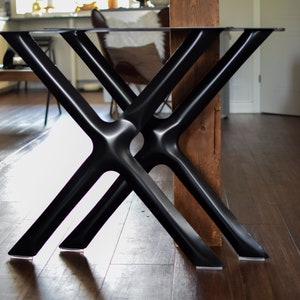 May include: Black metal table legs with a criss-cross design. The legs are attached to a glass tabletop and are resting on a wooden floor.