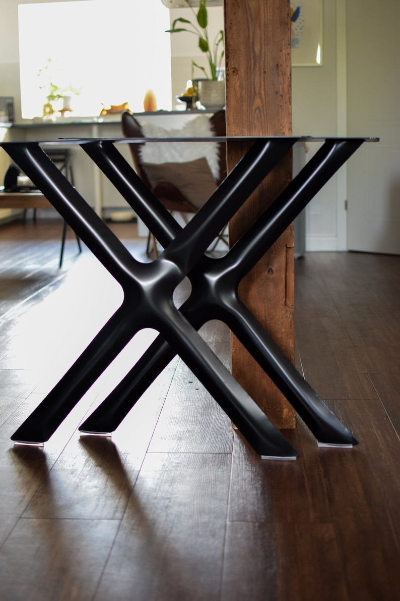 May include: Black metal table legs with a criss-cross design. The legs are attached to a glass tabletop and are resting on a wooden floor.