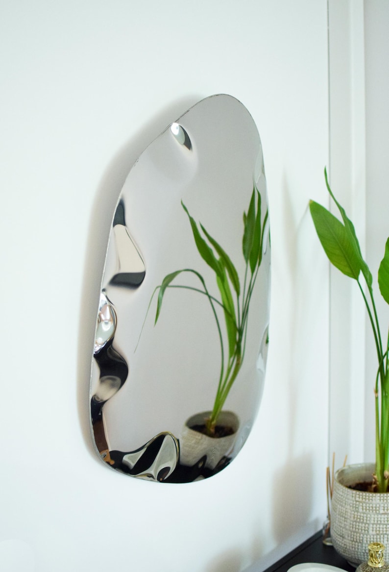 May include: A modern, oval-shaped mirror with a rippled surface hangs on a white wall. The mirror reflects a potted plant with green leaves. The mirror's unique design adds a contemporary touch to the room's decor.