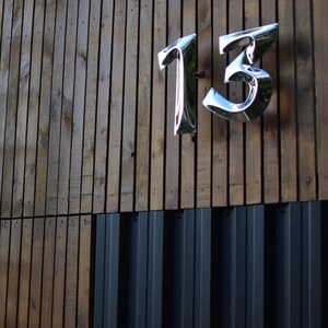 以下が含まれることがあります： 木製の壁に銀色の金属製の家の番号「13」が取り付けられている様子のクローズアップ。壁は濃い茶色の縦板で構成されている。