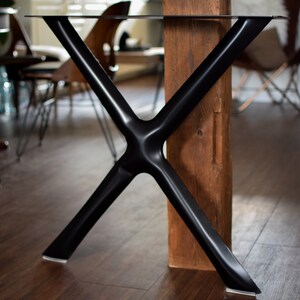 May include: Black metal table legs in an X shape, with a wood beam behind them. The legs are attached to a glass tabletop.