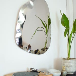 May include: A modern, irregularly shaped silver mirror hangs on a white wall. The mirror reflects a plant and pebbles. A turntable sits on a black table below the mirror, with a potted plant to the right.