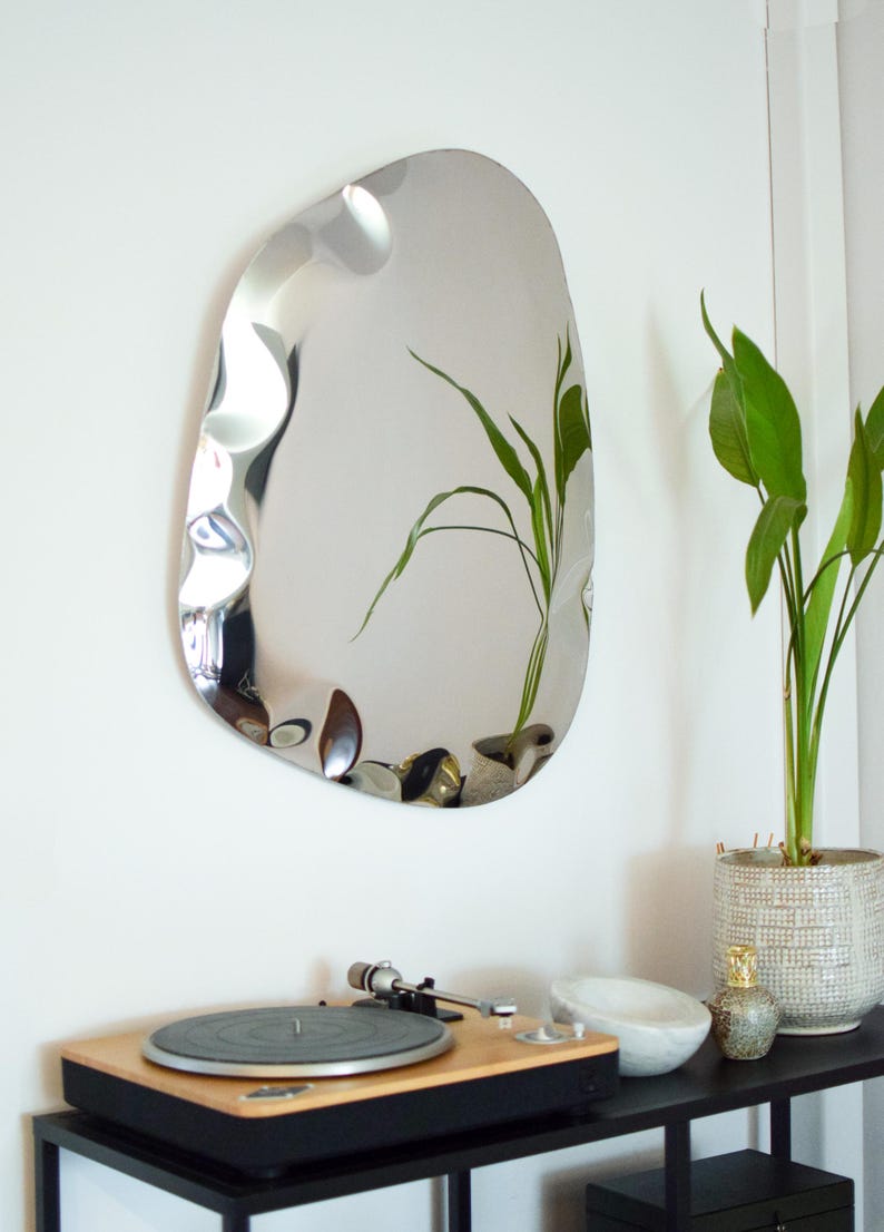 May include: A modern, irregularly shaped silver mirror hangs on a white wall. The mirror reflects a plant and pebbles. A turntable sits on a black table below the mirror, with a potted plant to the right.