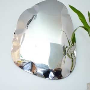 May include: A modern, irregularly shaped silver mirror hangs on a white wall. The mirror's surface reflects the room, with a distorted, wavy appearance. A green plant is visible to the right, and a turntable and decorative items are below.