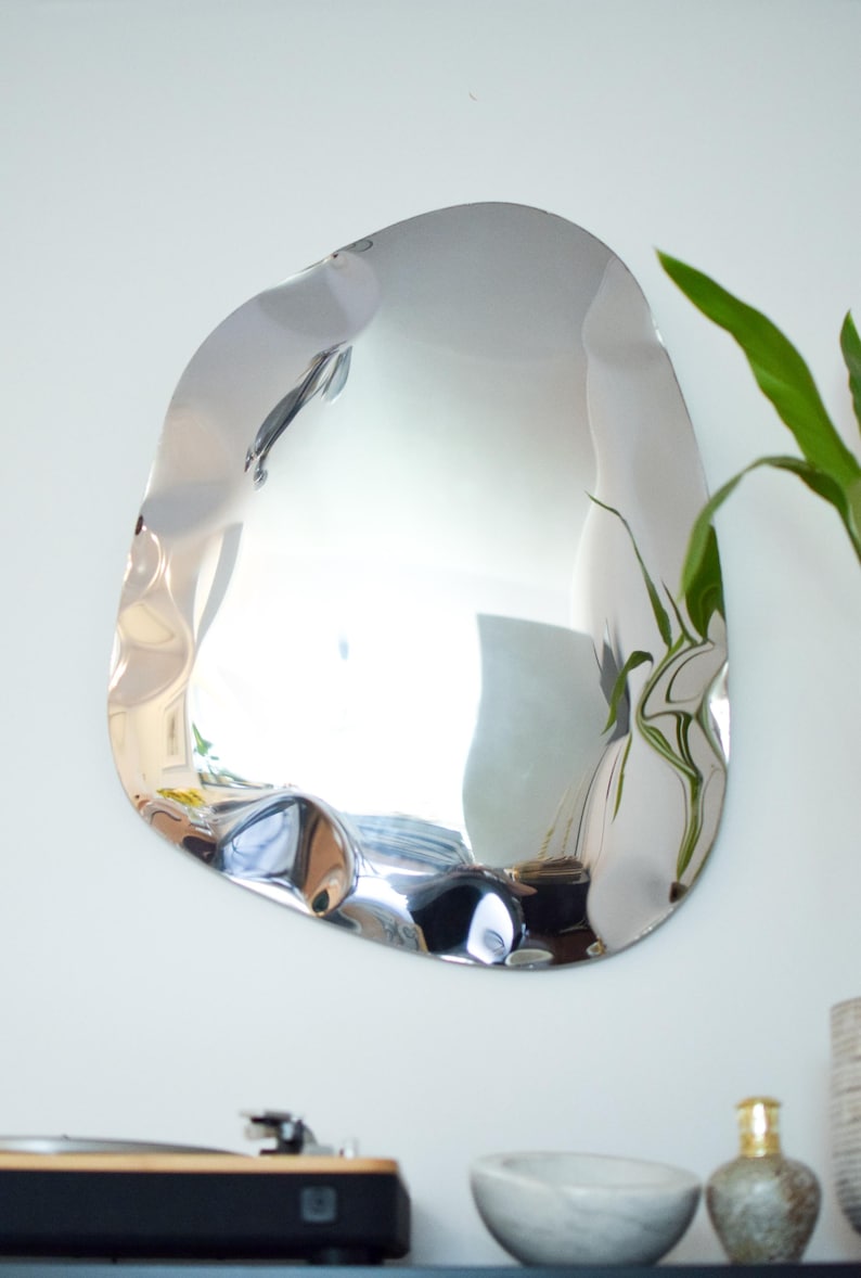 May include: A modern, irregularly shaped silver mirror hangs on a white wall. The mirror's surface reflects the room, with a distorted, wavy appearance. A green plant is visible to the right, and a turntable and decorative items are below.