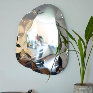 May include: A modern, irregularly shaped silver mirror hangs on a white wall. The mirror reflects the room, and a green plant in a textured pot sits nearby. A record player and a small bowl are visible below.