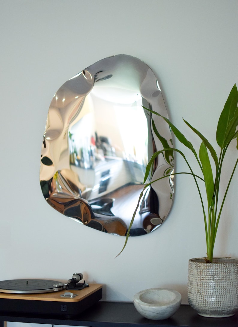 May include: A modern, irregularly shaped silver mirror hangs on a white wall. The mirror reflects the room, and a green plant in a textured pot sits nearby. A record player and a small bowl are visible below.