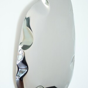 May include: A modern, irregularly shaped mirror with a polished, reflective silver surface. The mirror has a unique, organic form with subtle indentations, creating visual interest. It is mounted on a white wall, reflecting the surrounding environment.