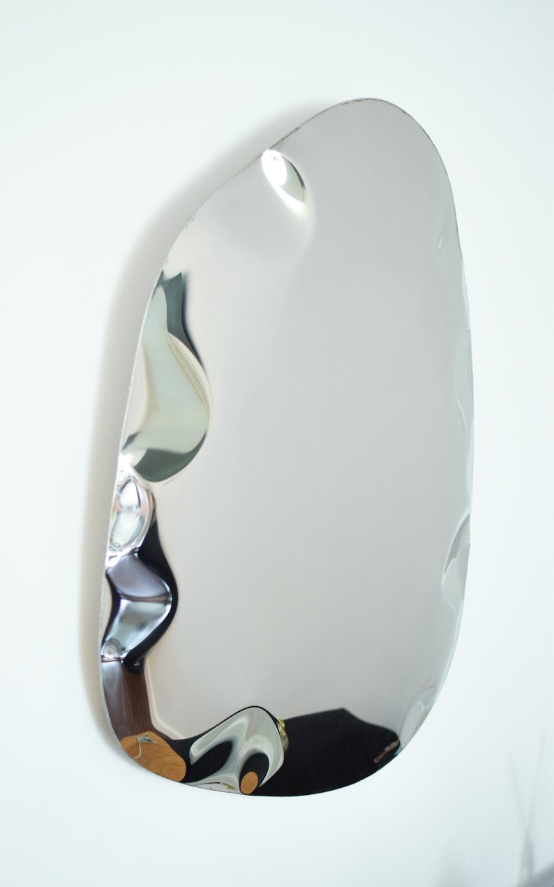 May include: A modern, irregularly shaped mirror with a polished, reflective silver surface. The mirror has a unique, organic form with subtle indentations, creating visual interest. It is mounted on a white wall, reflecting the surrounding environment.