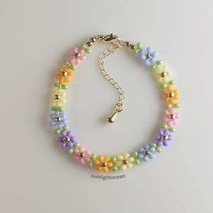 May include: A beaded bracelet featuring colorful flower designs in shades of pink, yellow, orange, green, blue, and purple. The bracelet has a gold-colored clasp and chain. The text "sunlightocean" is visible on the bottom.