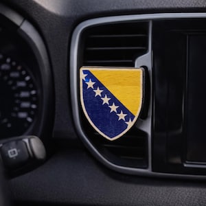 Könnte beinhalten: Dekoration aus Holz für die Lüftungsöffnung im Auto mit der Flagge von Bosnien und Herzegowina. Das schildförmige Emblem hat ein blaues Feld mit weißen Sternen und einen gelben oberen Bereich. Das Zubehör wird an der Lüftungsöffnung eines Autos befestigt.