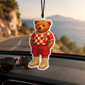 Peut inclure: Un désodorisant de voiture suspendu représentant un ours en peluche marron portant un maillot de football à damier rouge et blanc, un short rouge, des chaussettes beiges et des chaussures de football. L'ours a un petit emblème sur la poitrine.