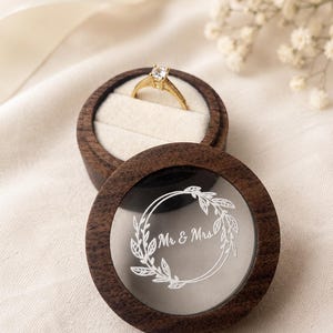 Op de afbeelding: Een ronde ringdoos van donker hout met een doorzichtig deksel met een witte gegraveerde bloemenkrans en de tekst Mr & Mrs. De doos staat open en toont een gouden ring met een enkele heldere steen op een crèmekleurig fluwelen kussentje.