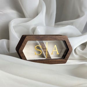 Pode incluir: Uma caixa de anel hexagonal de madeira com uma parte superior transparente. A caixa é marrom escuro com letras douradas no interior que dizem "S | A". O interior é forrado com um tecido de cor clara. A caixa está em um fundo branco drapeado.