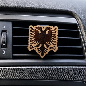 Può includere: Una clip deodorante per auto in legno con un design tagliato al laser di un'aquila bicipite. L'ornamento presenta una silhouette di aquila tinta in marrone scuro su uno sfondo di legno naturale più chiaro con dettagli intricati delle ali.