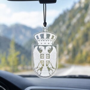 Könnte beinhalten: Ein hängendes Auto-Ornament aus klarem Acryl mit einem lasergravierten weißen Design des serbischen Wappens, bestehend aus einem Doppeladler, einem Schild mit Kreuz und vier Feuerstählen sowie einer Krone, aufgehängt an einer schwarzen Kordel.