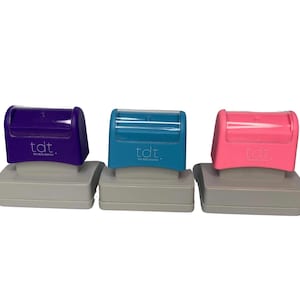 May include: Three self-inking stamps in purple, teal, and pink, each with a gray base. The stamps have a curved top with the text "tdt. the daily teacher." The stamps are designed for educational use.