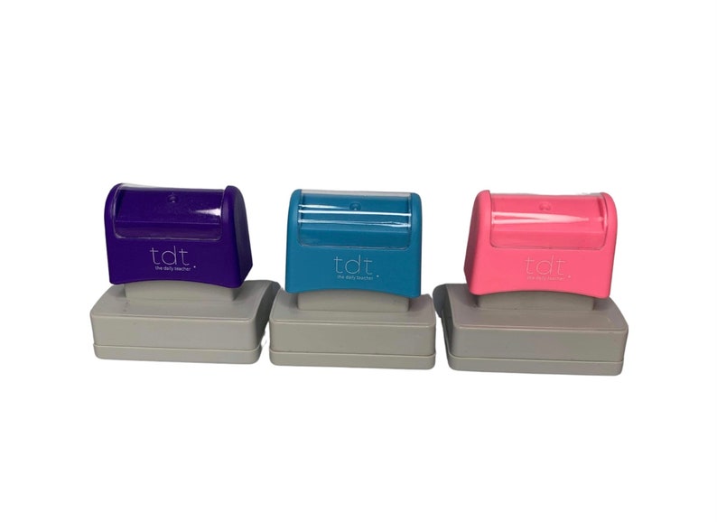May include: Three self-inking stamps in purple, teal, and pink, each with a gray base. The stamps have a curved top with the text "tdt. the daily teacher." The stamps are designed for educational use.