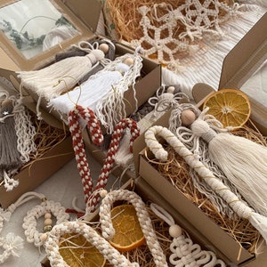 May include: A collection of handmade Christmas ornaments in a variety of styles, including angels, snowflakes, candy canes, and bells. The ornaments are made from natural materials like cotton and wood and are decorated with dried orange slices. The ornaments are packaged in brown boxes with straw filling.