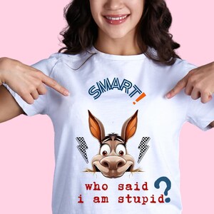 Peut inclure: Un t-shirt blanc à manches courtes avec un graphique de tête d'âne, deux éclairs et le texte "SMART!" en bleu et orange. Sous l'âne, le texte rouge indique "who said i am stupid?" accompagné d'un point d'interrogation bleu.
