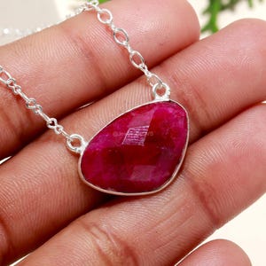 May include: A silver necklace featuring a large, irregularly shaped ruby gemstone pendant. The ruby is a deep, vibrant red color and is set in a silver bezel. The chain is a delicate silver link design. The necklace is displayed on a hand.