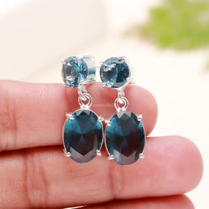 May include: A pair of silver earrings with two blue topaz gemstones. The top gemstone is round and the bottom gemstone is oval.
