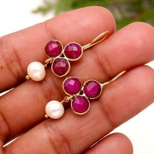 Puede incluir: Un par de pendientes dorados con grupos de tres piedras preciosas de color rojo intenso y una perla blanca. Las piedras están engastadas en un patrón circular, creando un diseño floral delicado.