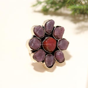 Puede incluir: Un anillo plateado con un diseño floral. El anillo está adornado con varias piedras preciosas en bruto de color rojo púrpura, con una sola piedra roja en el centro. El anillo se muestra sobre un fondo neutro.