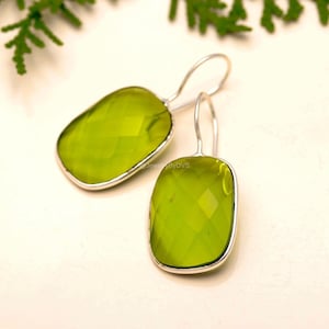 May include: A pair of lime green gemstone earrings with a faceted cut. The earrings have a rectangular shape with rounded corners and are set in a silver metal frame. The ear wires are also silver.