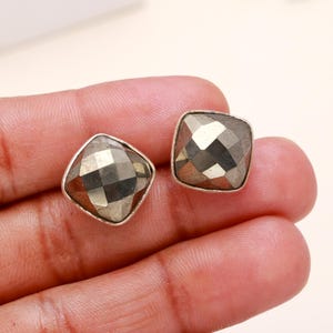 May include: A pair of square pyrite stud earrings. Each earring has a faceted, metallic gold-coloured pyrite stone set in a silver-toned bezel. The earrings are displayed against a neutral background.