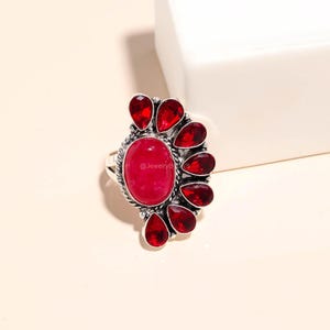 Può includere: Anello in argento con una grande pietra rossa ovale, circondata da pietre rosse più piccole a forma di goccia. L'anello ha una fascia dettagliata e una finitura lucida. Un gioiello di grande effetto.
