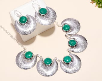 May include: A silver necklace and earrings set with green gemstone accents. The necklace features a chain with five crescent moon pendants, each with a green stone in the center. The earrings are also crescent moon shaped with green stones.
